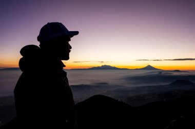 Siluetli bir adam dağın zirvesinde duruyor ve çarpıcı bir güneşin doğuşunu seyrediyor. Gökyüzü, uzak tepe ve vadileri aydınlatan koyu mordan parlak altına dönüşür..