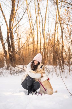 Sıcak kıyafetler giymiş bir kadın karlı bir ormanda köpeğini sevgiyle kucaklıyor. Kış manzarası, çıplak ağaçlar ve yumuşak güneş ışığı huzurlu ve rahat bir atmosfer yaratır..