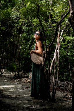 Uzun elbiseli ve güneş gözlüklü genç bir kadın Cozumel 'in tropik ormanının yemyeşil yeşilliklerinde gülümseyen bir ağaca yaslanıyor. Geleneksel bir hasır şapka taşıyor ve şık kıyafetine kültürel bir dokunuş katıyor..