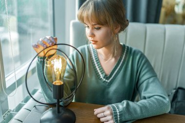 Sarışın bir kız, rahat bir kahve dükkanında, ahşap masadaki eski bir ampule nazikçe dokunuyor. Yumuşak ışık geniş bir pencereden süzülerek sıcak bir ortam yaratır.