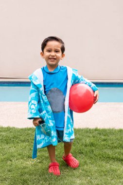 Mavi bornozlu ve kırmızı sandaletli neşeli bir çocuk yüzme havuzunun yanındaki çimlerin üzerinde duruyor, kırmızı bir top tutuyor ve gülümsüyor, kaygısız ve neşeli bir anı yakalıyor..
