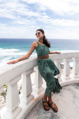 Karayip Denizi 'ne ve engebeli Cozumel sahil şeridine bakan beyaz bir balkona yaslanan neşeli bir kadın. O, güneş gözlükleriyle tamamlanmış, dalgalı yeşil bir elbise giydi..