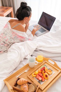 Yukardan bakıldığında, hafta sonu evde dinlenirken sağlıklı bir kahvaltıyla rahat bir yatakta yatarken dizüstü bilgisayar kullanan genç bir kadın görülüyor.