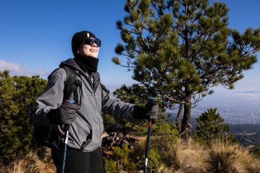 Yürüyüş direkleri ile coşkulu bir yürüyüşçü dağda güneşli bir kış gününün tadını çıkarıyor. Ağaçlarla çevrili, kışlık giysiler giyiyor ve açık hava macerasını kucaklıyor..