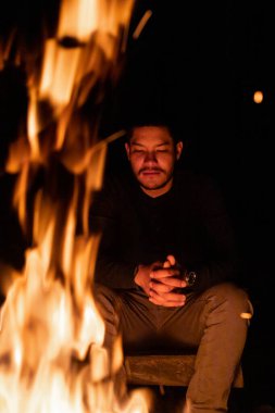 Bir adam kamp ateşinin yanında sessizce oturur, alevler düşünceli ifadesini aydınlatır. Onu çevreleyen gece sakin. Ormanın huzurlu atmosferini ve iç dünyasını canlandırıyor..