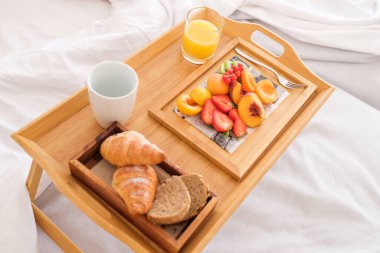 Tahta tepsinin tepsisinden lezzetli kruvasanlar ve taze meyve ve içeceklerle servis edilen ekmek dilimleri sabah beyaz yatakta.