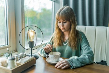 Genç sarışın bir kadın şık bir kafede huzurlu bir anın tadını çıkarırken kahvesini karıştırıyor. Sıcak ışıklandırma ve rahat oturma rahatlama için mükemmel bir ortam yaratır.