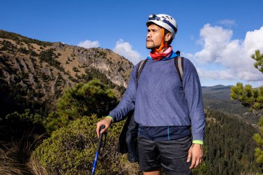 Kask ve far takmış bir yürüyüşçü, bastonla ayakta duruyor, manzaralı dağ manzarasına bakıyor. Ağaçlar ve bulutlar arka planda, açık hava macerasını gösteriyor..