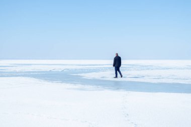 Koyu renk ceketli yalnız bir adam, açık mavi gökyüzünün altında, engin, donmuş bir gölde yürüyor. Kış aylarında yalnızlık ve sükuneti çağrıştırıyor..