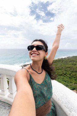 Neşeli bir kadın Cozumel, Meksika 'da manzaralı bir manzarada selfie çekerken sevinçle kolunu kaldırıyor. Güneş gözlüğü takıyor, desenli bir elbise ve bereketli kıyı manzarasıyla çevrili..