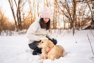 Kışlık ceket giyen ve örgü şapkalı bir kadın karlı bir orman manzarasında Golden retriever 'ının yanında diz çöker. Güneş arka planda batar, ılık bir parıltı yayar..