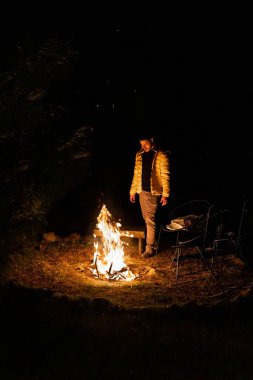 Bir adam uzak bir ormanda gece gökyüzünün altında çatırdayan bir kamp ateşinin yanında duruyor. Yangının sıcaklığı, doğayla iç içe, huzurlu ve huzurlu bir akşam atmosferi yaratır..