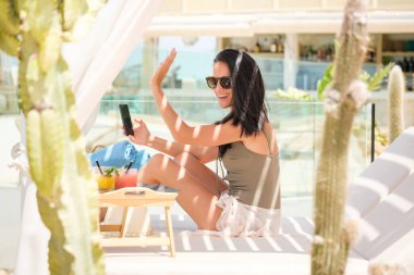 Güneş gözlüğü dalgaları içinde neşeli bir kadın telefonuyla görüntülü sohbet ederken, şık bir otel kabininde oturmuş tropikal içecekler, kaktüs bitkileri ve çarpıcı güneşli bir manzarası olan.