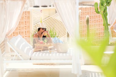 Güneş gözlüklü ve hasır şapkalı şık bir kadın lüks bir tatil köyünde yemyeşil, modern dekor ve sıcak güneş ışığıyla çevrili bir yerde tropikal bir içkiyi yudumlarken özel bir kabinde dinleniyor..