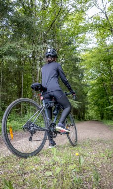 Siyah kıyafetli ve kasklı bisikletçi baharda ormanda bisiklet sürüyor. Bereketli yeşilliklerle çevrili sahne, sakin bir doğal ortamda açık hava macerasını yakalıyor..