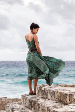 Cozumel 'de okyanusa bakan kayalara bakan, bulutlu bir günde yeşil bir elbise giyen bir kadının arkası..