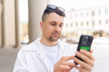 Beyaz elbiseli bir erkek işadamı, kentsel bir ortamda dikilirken dikkatle akıllı telefonuna odaklanır..