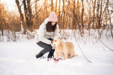 Karlı bir ormanda altın av köpeğinin yanında diz çöken bir kadın. Pembe bir bere ve kışlık bir ceket giyer. Sadık arkadaşıyla birlikte sakin bir kış gününün tadını çıkarır..