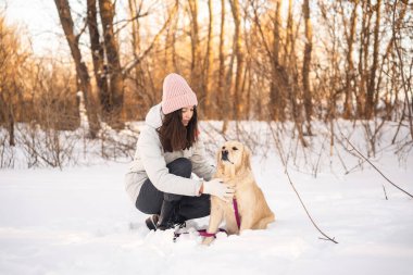 Pembe şapkalı bir kadın, karlı bir ormanda altın köpeğinin yanında diz çöker. Köpek bir koşum takımı takıyor. Sahne, çıplak ağaçların arasında huzurlu bir kış anını yakalıyor..