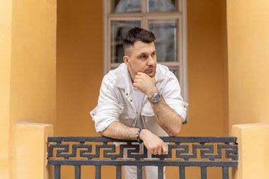 Zarif beyaz elbiseli genç bir işadamı süslü bir balkon korkuluğuna yaslanırken, yumuşak sarı bir binanın arka planına yaslanıyor..