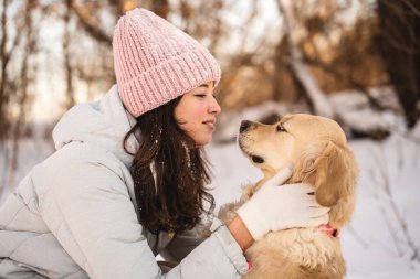 Pembe şapkalı ve beyaz önlüklü bir kadın karlı bir ormanda köpeğini kucaklıyor ve yakın bağlarını parlaklaştırıyor. Etraflarına kar taneleri yavaşça düşer ve sakin bir kış ortamı yaratır..