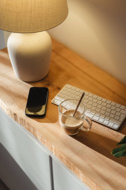 Sıcak bir lamba, bir bardak temiz kahve, bir akıllı telefon ve ahşap bir masanın üzerinde bir klavye içeren rahat bir çalışma alanı, huzurlu ve üretken bir atmosfer yaratıyor..