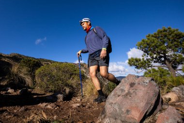 Yürüyüş direkleri ve miğferiyle donatılmış bir yürüyüşçü açık mavi gökyüzü altında engebeli bir dağ yolunda yürüyor. Yeşillik ve kayalık arazi çarpıcı bir doğal zemin oluşturur..