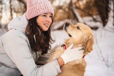 Kışın giyinmiş bir kadın, karlı bir ormanda bir golden retriever 'ı kucaklıyor. Her ikisi de, aralarındaki bağın altını çizerek, sakin kış manzarasında sıcaklığı ve mutluluğu ifade ediyor..