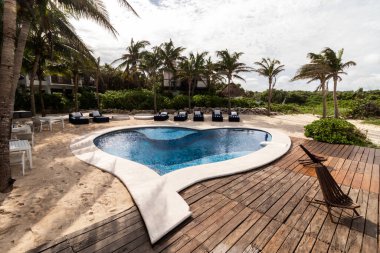 Palmiye ağaçları ve plaj şezlonglarıyla çevrili berrak mavi suyu olan sakin bir havuz, yemyeşil ve kumlu sahillerin zeminine kurulmuş..