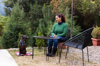 Yeşil kazaklı bir kadın bahçe masasında oturuyor ve köpeğiyle huzurlu bir anı paylaşıyor. Bereketli yeşilliklerle çevrili sahne, sükuneti ve arkadaşlığı yakalıyor..