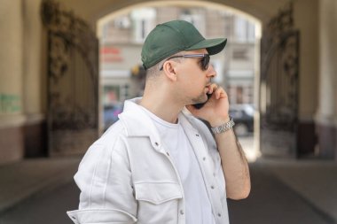 Beyaz elbiseli ve yeşil şapkalı bir işadamının yan görüntüsü meşgul bir şehir caddesinde dikilirken cep telefonuyla sohbet ediyor..