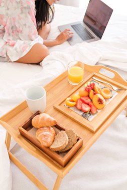 Anonim kadın seyahat blogcusu dizüstü bilgisayarını kullanarak sabah modern otellerde sunulan lezzetli sağlıklı kahvaltıyla ahşap tepsinin yanında oturuyor.