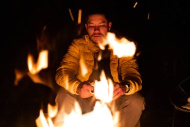 Sarı ceketli bir insan, gece ormanda canlı bir kamp ateşinin yanında sıcak bir içecekten zevk alır. Sahne, dışarıdaki sıcaklığı, rahatlığı ve sükuneti yansıtıyor..