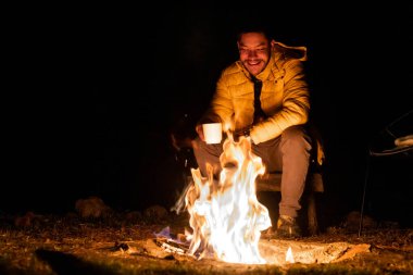 Sarı ceketli bir insan, ormanda serin bir gecede kamp ateşinin yanında sıcak bir içkinin tadını çıkarır. Alevler karanlık zemine karşı dans ediyor, rahat bir açık hava ortamı yaratıyor..