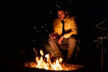 Sarı ceketli bir insan, ormanda yıldızlı bir gökyüzünün altında kamp ateşinin yanında içmekten zevk alır. Ateşin sıcak parıltısı rahat ve huzurlu bir atmosfer yaratır..