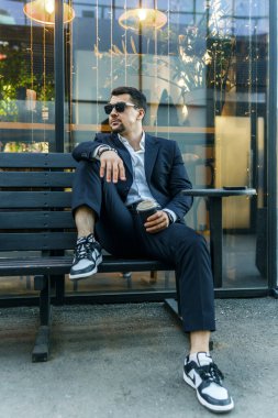 Takım elbiseli şık bir işadamı dışarıdaki ahşap bir bankta, elinde kahve fincanıyla rahatça oturur. Rahat ve kendinden emin görünüyor, modern şehir hayatını somutlaştırıyor.