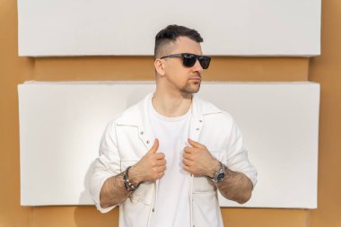 Beyaz şık giyinmiş ve güneş gözlüğü takmış kendine güvenen bir erkek işadamı bej renkli yumuşak bir arka plana karşı duruyor..
