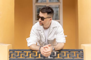 Kendine güvenen beyaz elbiseli ve güneş gözlüklü bir erkek işadamı şehir ortamında bir korkuluğa yaslanıyor ve modern profesyonel bir görünüm sergiliyor..