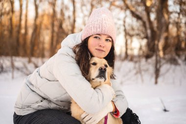 Kışın giyinmiş bir kadın, karlı bir yürüyüş sırasında golden retriever 'ına sarılır. Sakin kış ormanı arka planında soğuk mevsimde sıcaklık ve dostluk yayılıyor..