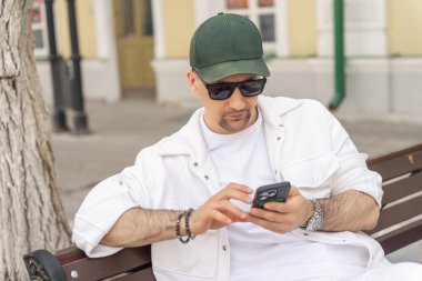 Şık beyaz giysiler içinde odaklanmış bir adam şehir bankında oturmuş, akıllı telefonuna bakıyor..