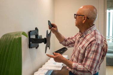 Yaşlı bir adam, iyi aydınlatılmış bir odadaki çeşitli aletleri kullanarak, bir televizyon desteğini güvenli bir şekilde duvara monte etmeye odaklanır..