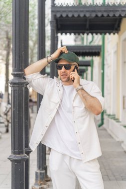 Beyaz takım elbiseli şık bir erkek işadamı büyüleyici bir şehir ortamında akıllı telefonuyla konuşurken klasik bir sokak lambasına yaslanıyor..