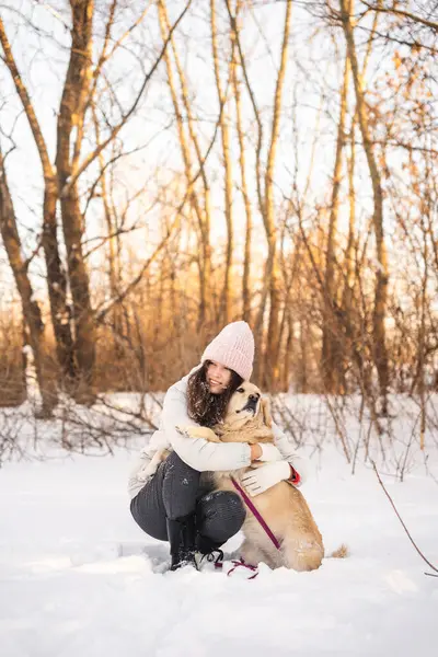 Sıcak kıyafetler giymiş bir kadın karlı bir ormanda köpeğini sevgiyle kucaklıyor. Kış manzarası, çıplak ağaçlar ve yumuşak güneş ışığı huzurlu ve rahat bir atmosfer yaratır..
