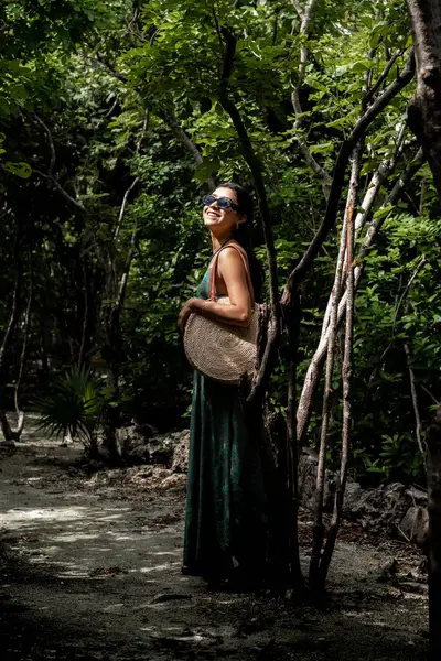Uzun elbiseli ve güneş gözlüklü genç bir kadın Cozumel 'in tropik ormanının yemyeşil yeşilliklerinde gülümseyen bir ağaca yaslanıyor. Geleneksel bir hasır şapka taşıyor ve şık kıyafetine kültürel bir dokunuş katıyor..