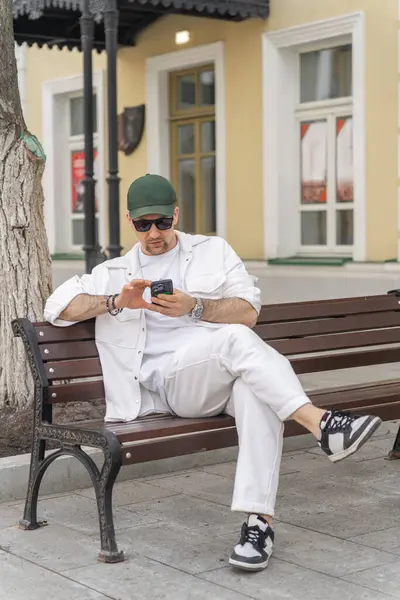 Beyaz elbiseli bir erkek işadamı bir ağacın altındaki bankta otururken akıllı telefonuna odaklanır..