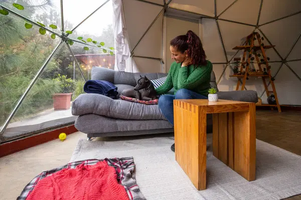 Bir kadın, köpeği eşliğinde modern bir glamping 'in içinde bir kanepede dinlenir. Konforlu odada ahşap bir masa, bitkiler ve doğal ışık var..