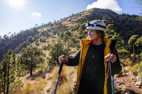 Yürüyüş yapan kadın miğfer, elinde sırıklar, güneşli bir dağ manzarasının tadını çıkarırken gülümsemeler, macera ve doğa keşifleri..