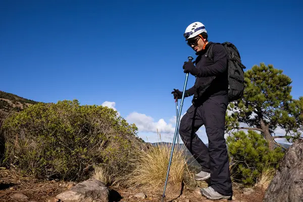 Açık hava kıyafeti giyen bir yürüyüşçü ve yeşil ve açık mavi gökyüzü ile çevrili kayalık bir dağ yolunda yürüyüş direkleri ile yürüyen bir kask. Macera ve doğa kavramı.