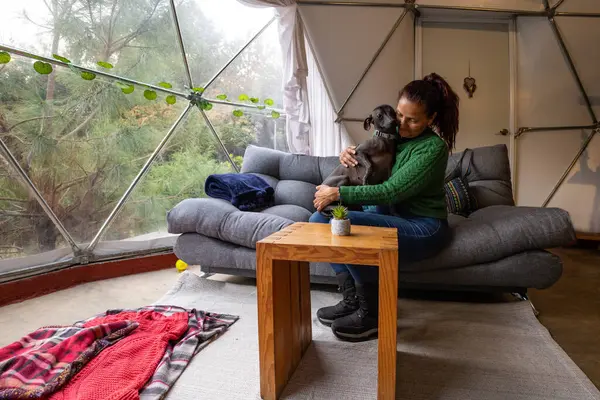 Bir kadın köpeğiyle modern bir glampingin içinde huzurlu bir anın tadını çıkarır. İç kısımda rahat bir kanepe, ahşap masa ve doğa manzarası sunan geniş pencereler yer alıyor..