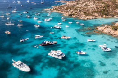 Lüks yatların ve teknelerin, seyrek bitki örtüsü olan kayalık bir sahil şeridi yakınındaki Akdeniz 'in berrak turkuaz sularında gezindiği hava manzarası. Üst dron görüntüsü. İtalya, Sardunya 'da yaz tatili